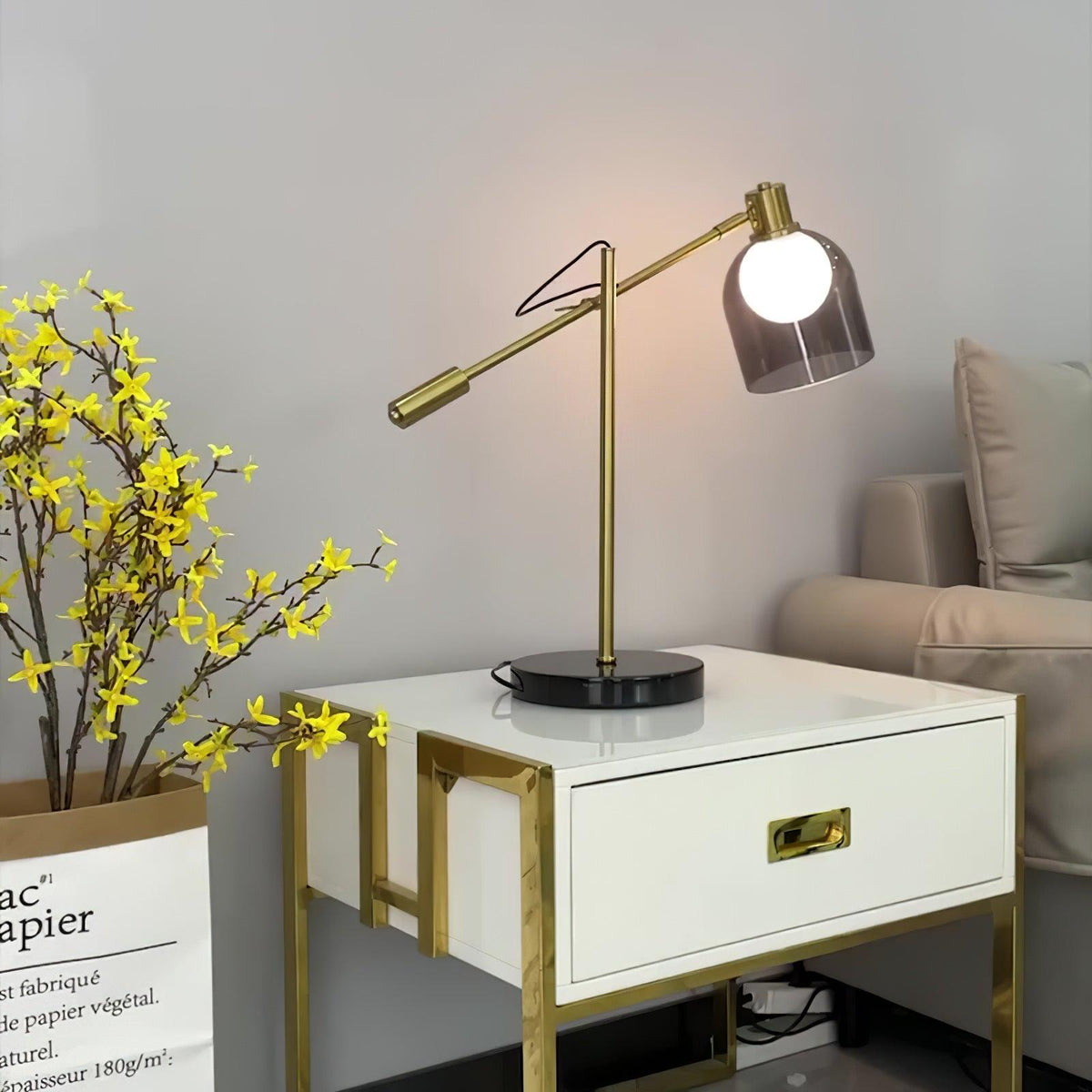 Clérachic brass table lamp lit and glowing on a white nightstand with brass drawer trim, beside a vase of yellow flowers.