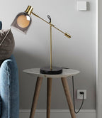 Against a grey wall, a Clérachic brass table lamp sits on a round marble-topped side table, denim fabric and a socket at the frame's edge.