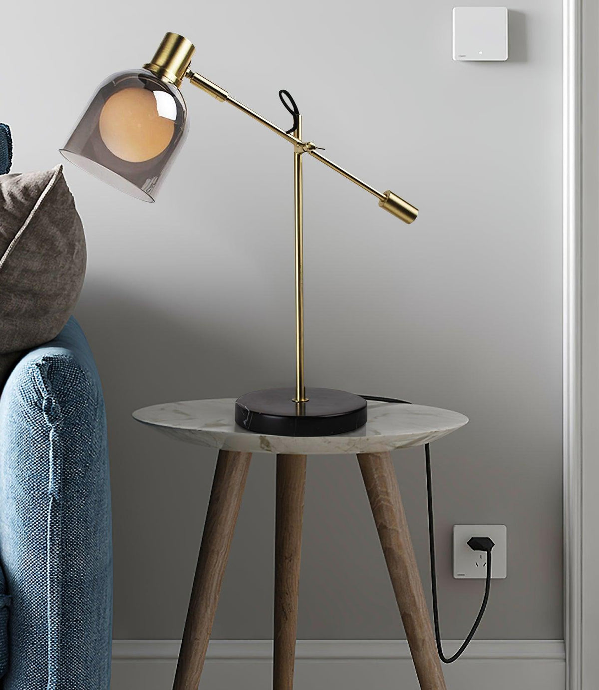 Against a grey wall, a Clérachic brass table lamp sits on a round marble-topped side table, denim fabric and a socket at the frame's edge.