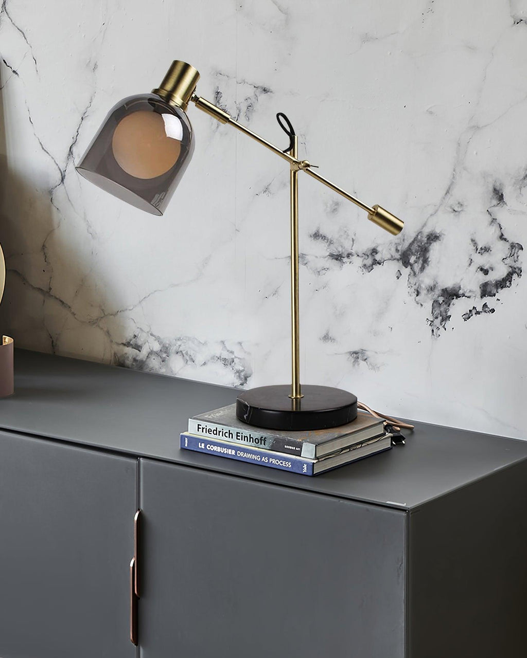 Clérachic brass table lamp set on a stack of books atop a dark grey cabinet, backed by a marble-tiled wall.