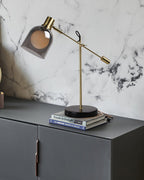 Clérachic brass table lamp set on a stack of books atop a dark grey cabinet, backed by a marble-tiled wall.