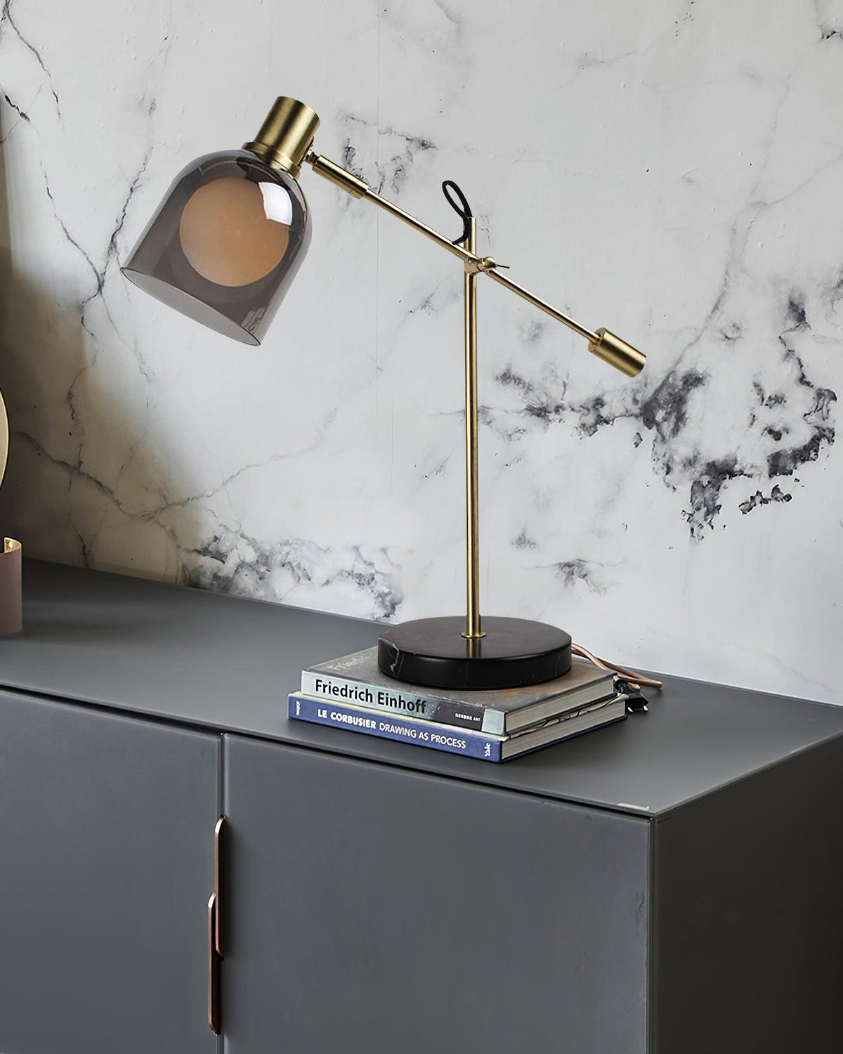 Clérachic brass table lamp set on a stack of books atop a dark grey cabinet, backed by a marble-tiled wall.