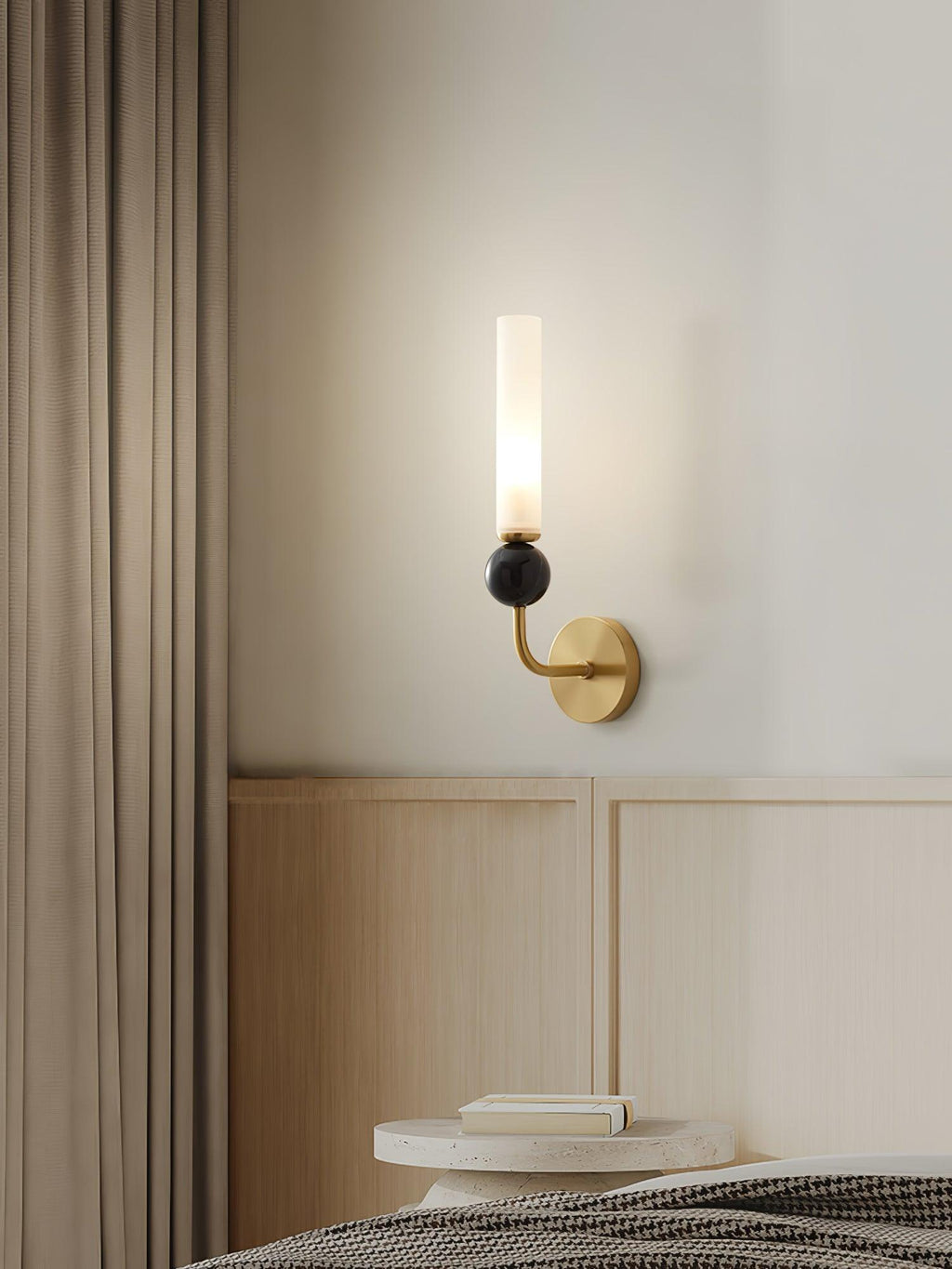 A Clérachic black wall sconce lights a bedside table stacked with books, set against a dark curtain beside the headboard.