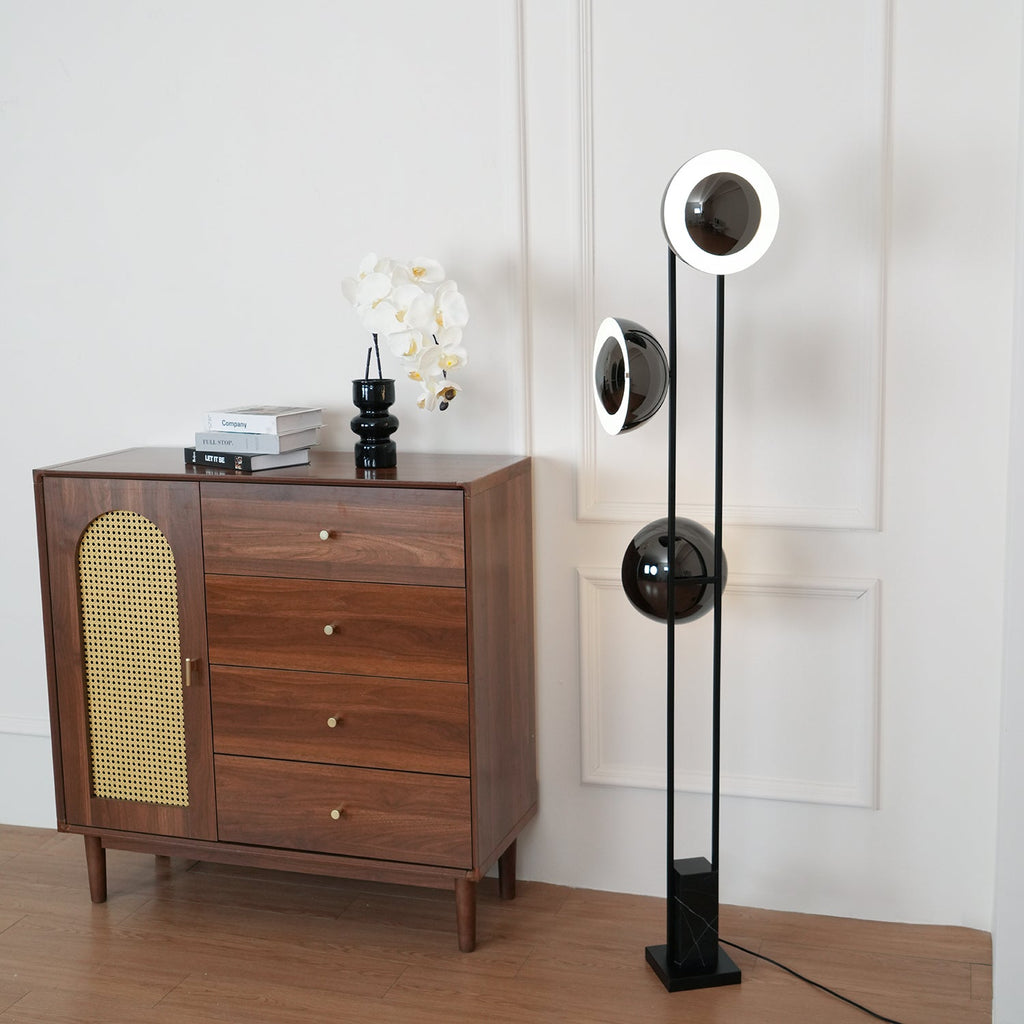 Clérachic floor lamp with its top ring lit, angled beside a wooden chest of drawers with an orchid on top.
