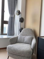 Partial view of the Clérachic floor lamp beside a curtained window and a grey tweed armchair in a quiet corner.