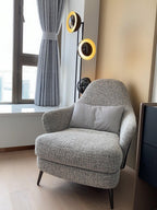 Top ring of the Clérachic floor lamp lit gold beside a curtained window and a grey tweed armchair in a bedroom corner.