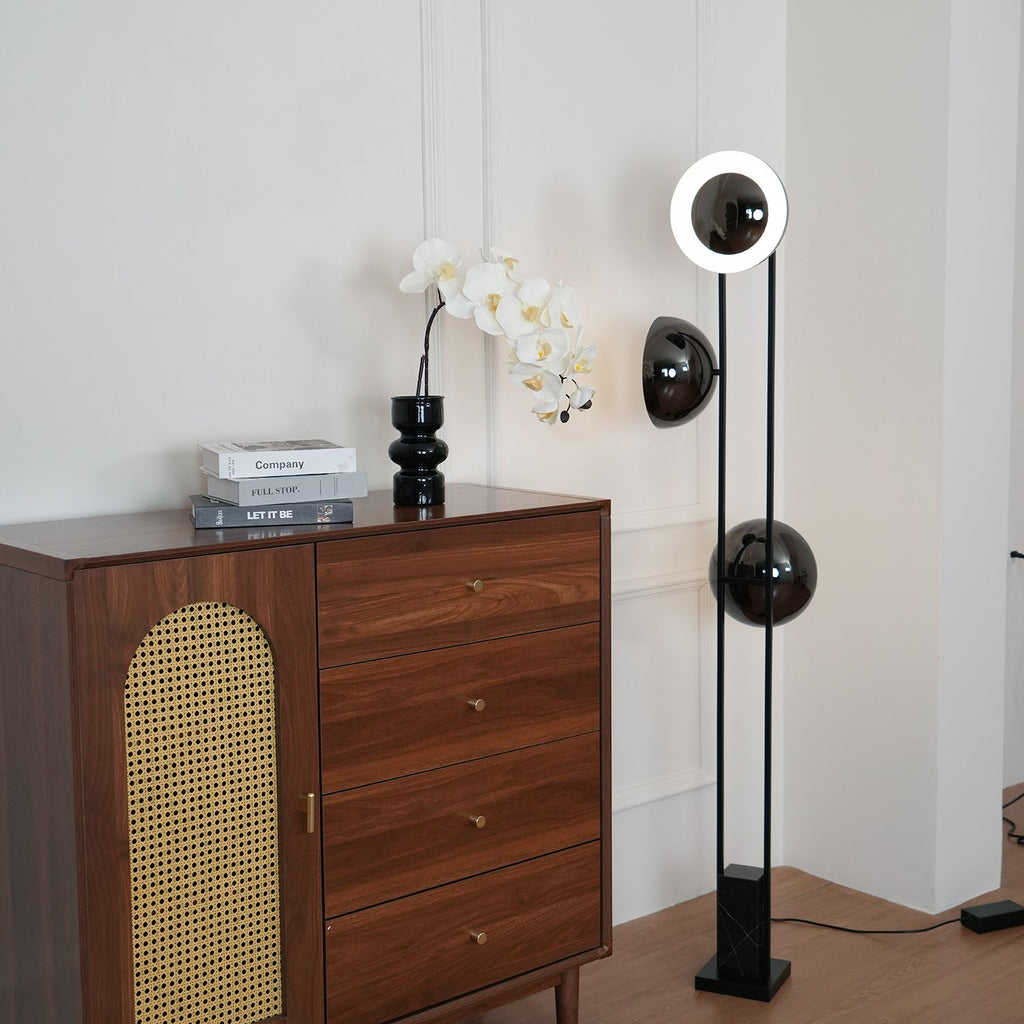 Slim black floor lamp by Clérachic, top ring glowing white, positioned beside a walnut and rattan chest of drawers.