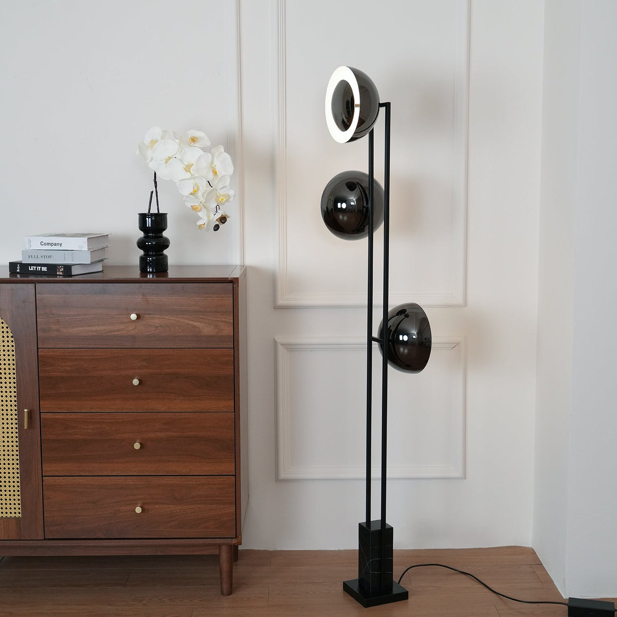 Clérachic floor lamp with its top ring lit, angled beside a wooden chest of drawers with an orchid in a black vase.