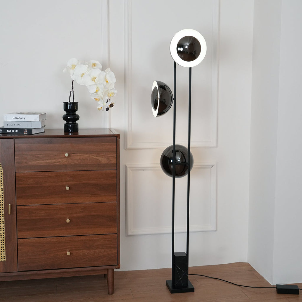Black three-light floor lamp from Clérachic glowing beside a walnut chest of drawers topped with an orchid and books.