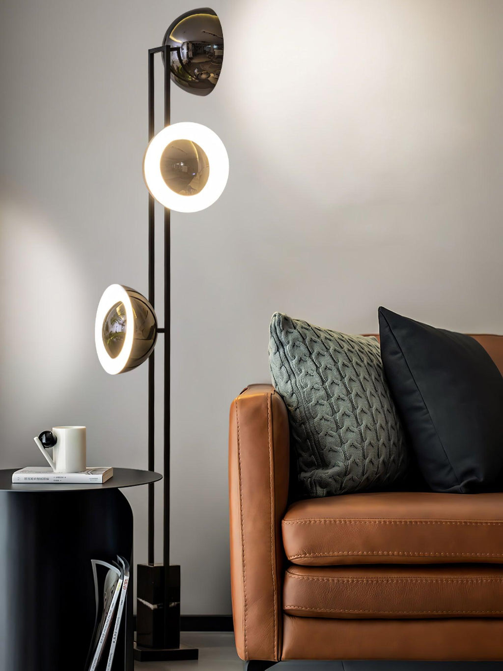 Middle and lower heads of the Clérachic floor lamp glowing beside a round side table with a mug and books on a tan sofa.