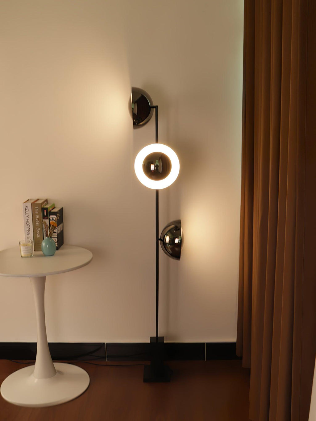 Clérachic floor lamp with its lower ring lit warm white, beside a round table stacked with books near a curtain.