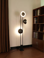 Clérachic floor lamp with its top and lower rings lit, standing in front of a wooden bookshelf and curtain.