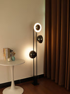 Clérachic floor lamp with its top ring lit, standing beside a round side table with books near a curtained wall.