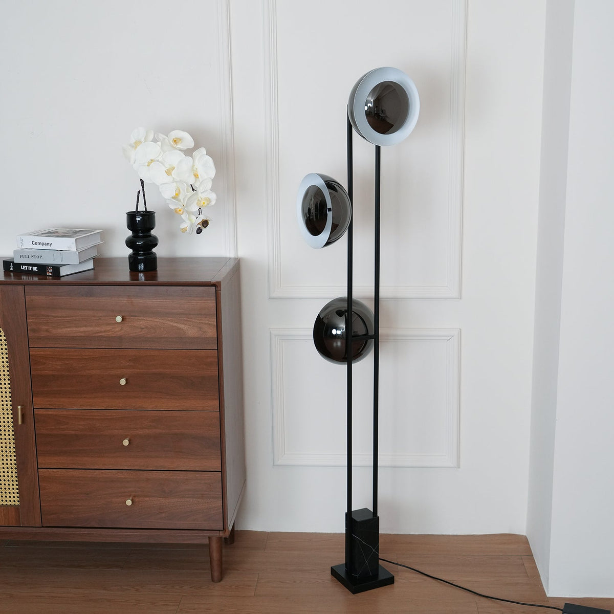 Clérachic floor lamp with its top ring lit pale white, standing beside a rattan-fronted chest topped with an orchid.