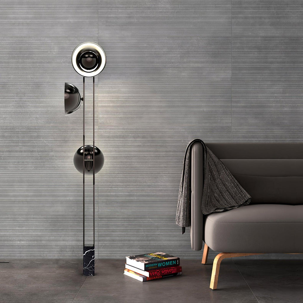 Clérachic three-light floor lamp glowing against a grey wall beside a sofa with a folded throw and a stack of books.
