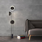 Clérachic three-light floor lamp glowing against a grey wall beside a sofa with a folded throw and a stack of books.
