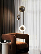 Clérachic floor lamp with its lower ring lit warm white, standing beside a burnt-orange chair and a sheer curtain.