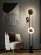 Clérachic floor lamp with its lower ring lit warm white, standing beside a taupe armchair and an abstract painting.