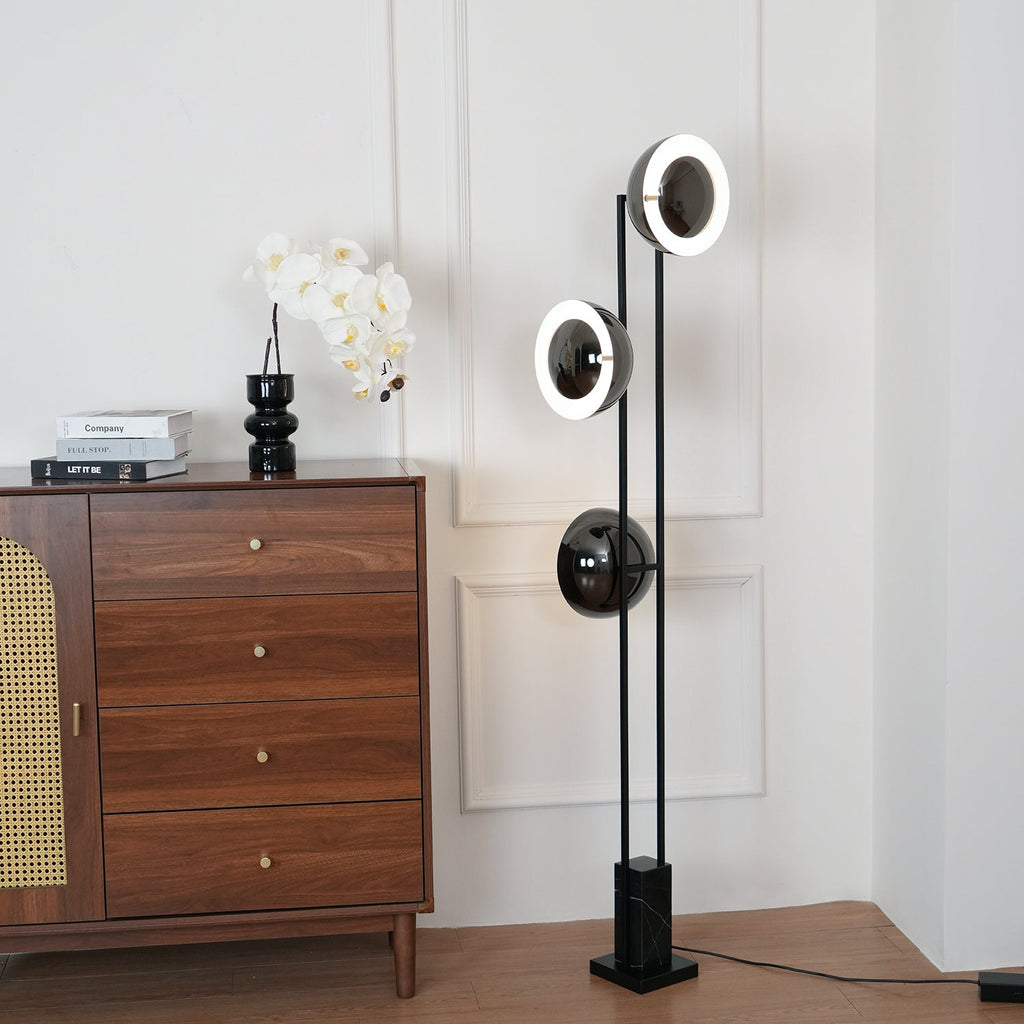 Clérachic three-light floor lamp with its upper ring lit, standing beside a wooden chest topped with an orchid and books.