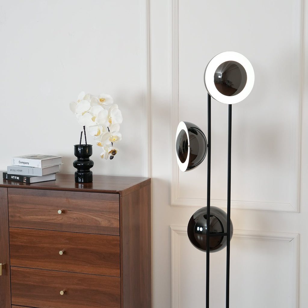 Clérachic floor lamp with its top ring lit white, standing beside a chest of drawers dressed with an orchid and books.