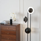 Clérachic floor lamp with its top ring lit white, standing beside a chest of drawers dressed with an orchid and books.