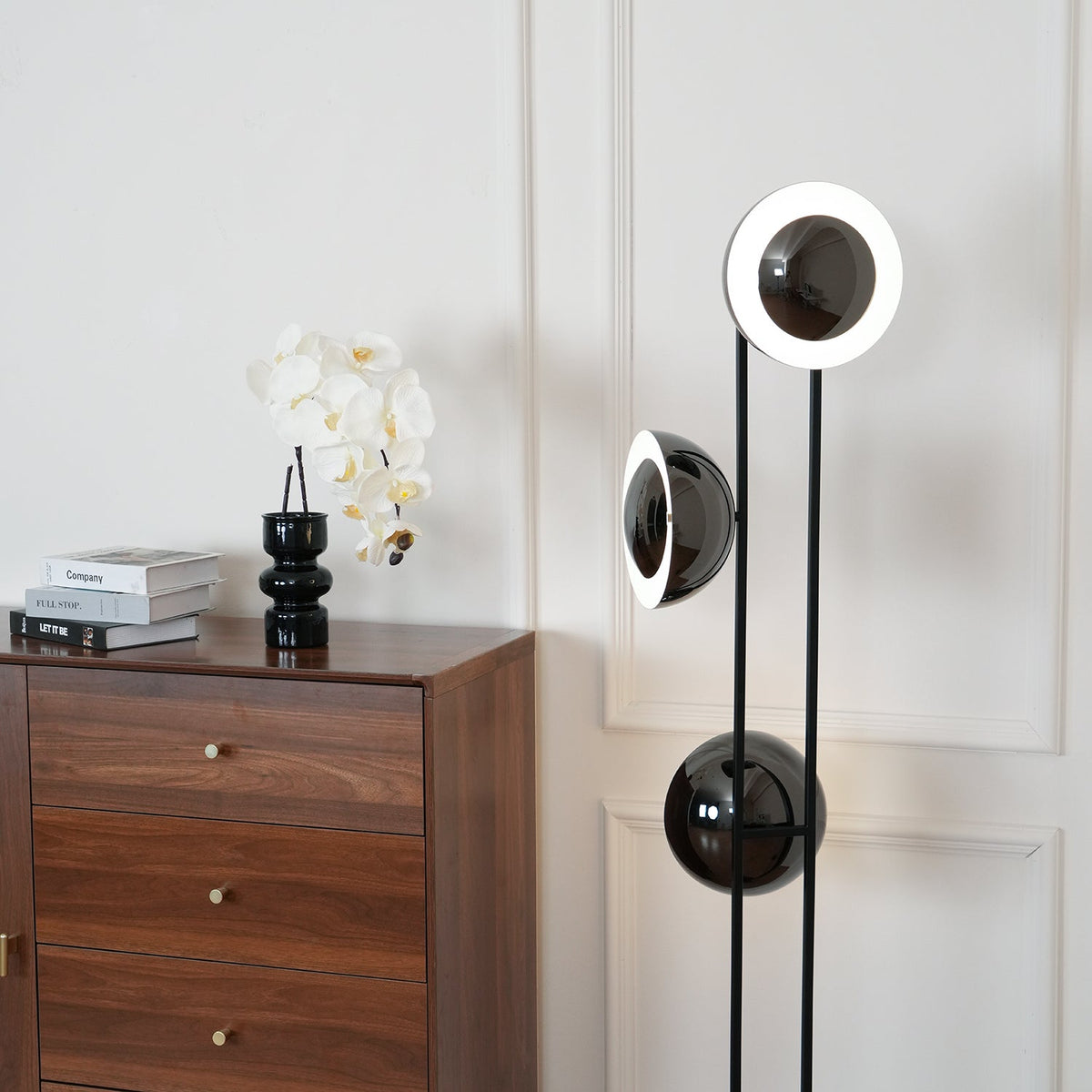 Clérachic floor lamp with its top ring lit white, standing beside a chest of drawers dressed with an orchid and books.