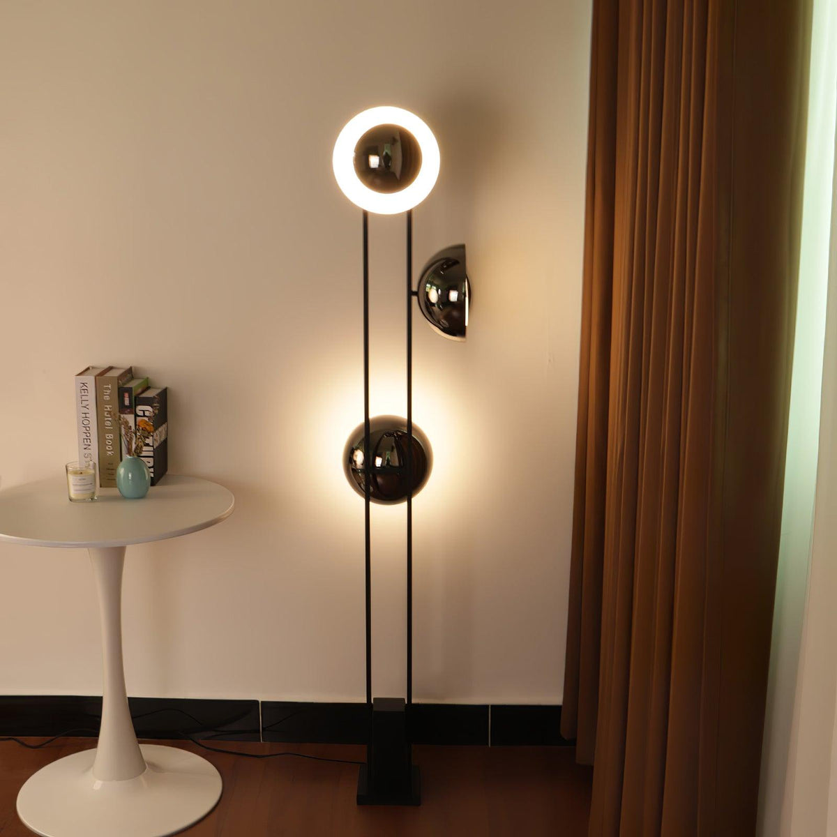Clérachic floor lamp with its top ring lit, positioned near a round white table and curtain in a warm-toned room.