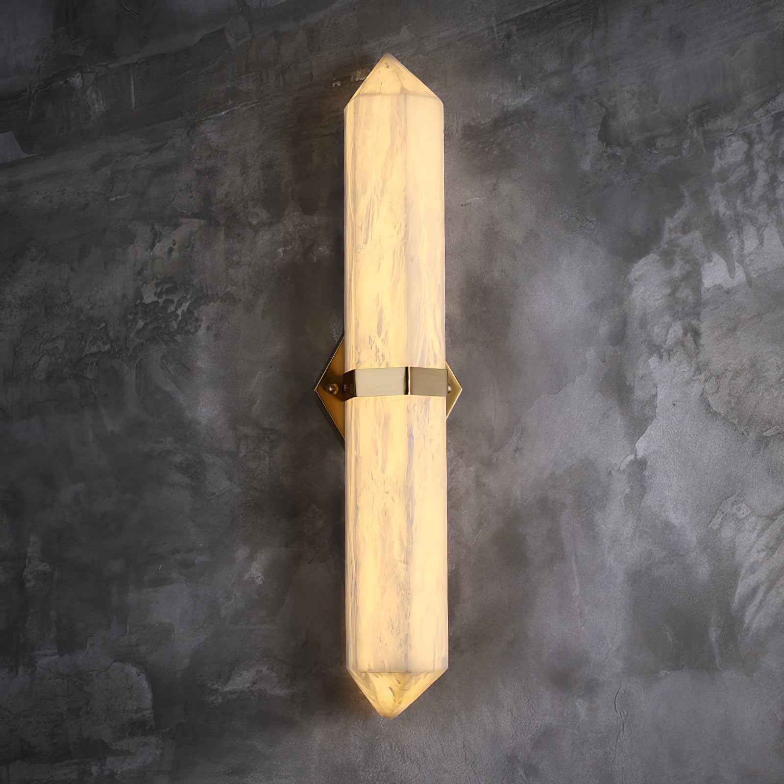 The brass-band sconce lit on grey concrete, seen full length from top point to bottom point.