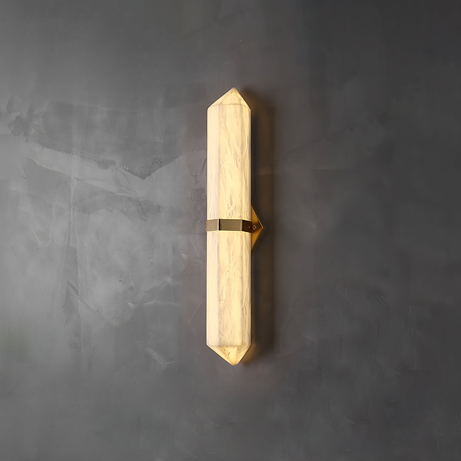 Faceted alabaster obelisk sconce lit on a grey concrete wall, held by a slim brass band.