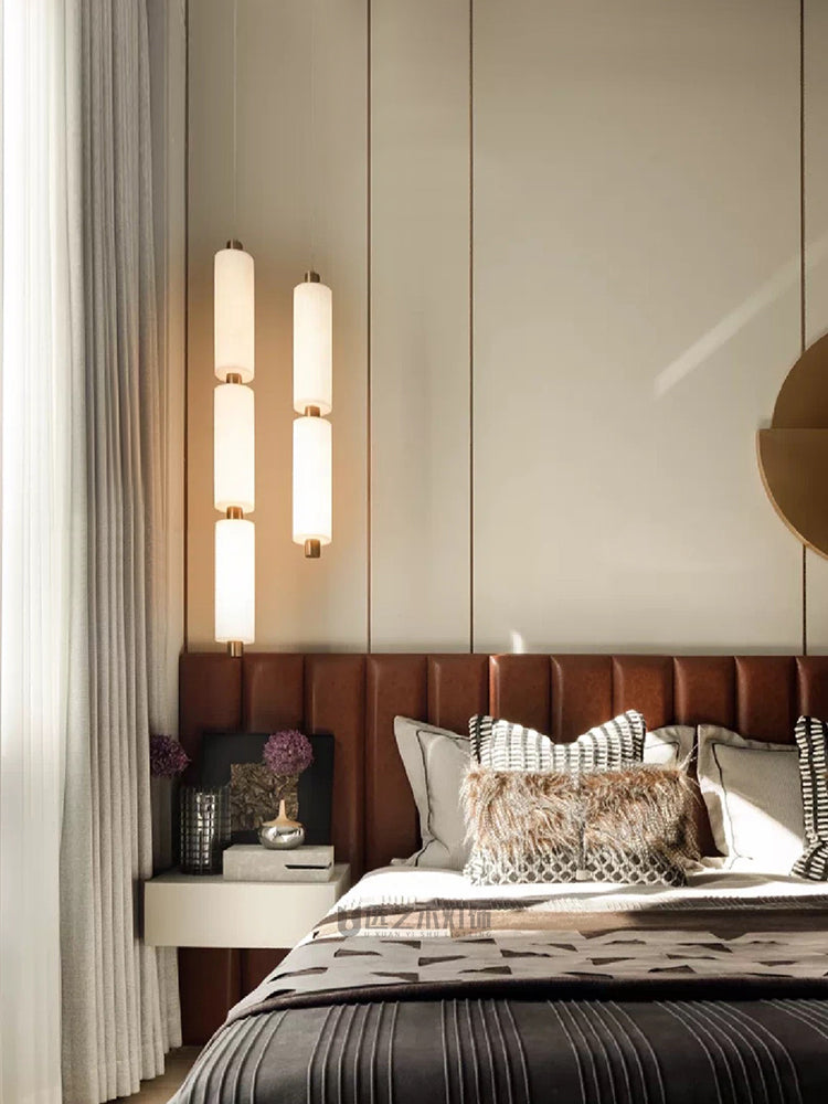 Two Zovarion alabaster pendants lit at staggered drops beside a tan leather headboard in a bedroom.