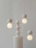 Grey stone-textured pendants from this Clérachic collection hang beside a stacked stone pedestal on a plain backdrop.