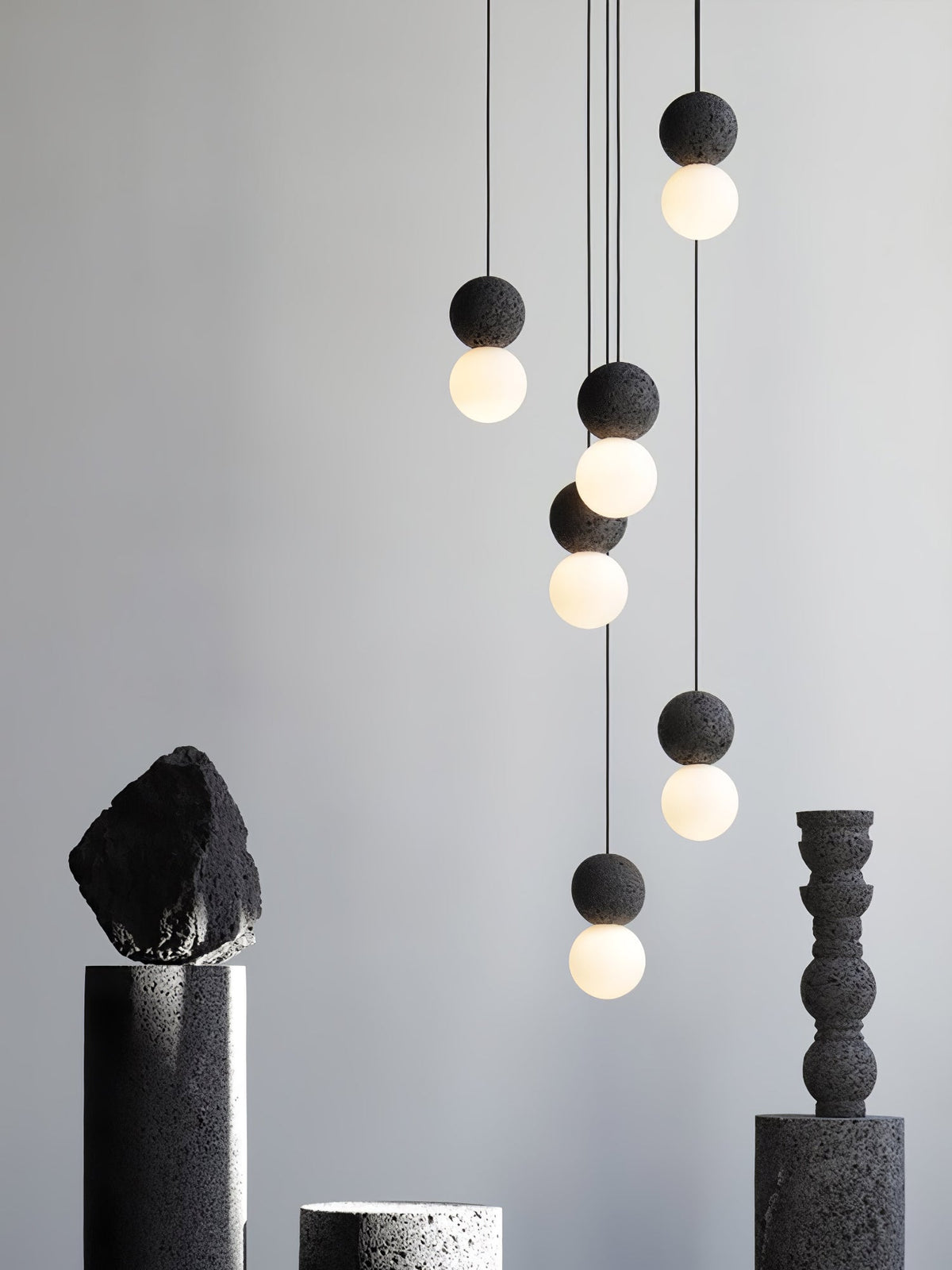 Several dark sphere pendants from this Clérachic collection hang above dark stone and ceramic pedestals on a plain grey backdrop.