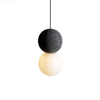 Close view of this Clérachic pendant, a large dark stone-textured sphere above a glowing opal glass globe, on a black cord.