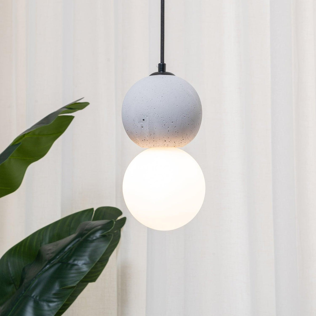 Close view of a light grey stone pendant with an opal glass globe, this Clérachic light hanging near sheer curtains and a leafy plant.