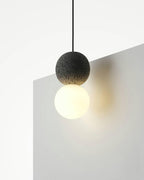 Close view of this Clérachic pendant, a dark stone-textured sphere above a warm-lit opal glass globe, against a plain wall corner.