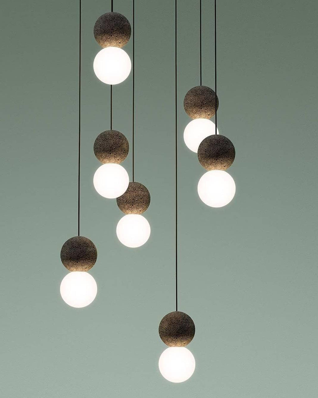 Several Clérachic pendants cluster together against a sage-green wall, their dark spheres and glass globes staggered at different heights.