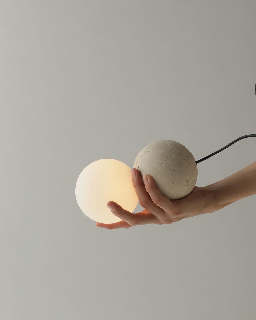 A hand holds this Clérachic pendant close, its opal glass globe softly lit beneath a dark stone-textured sphere.