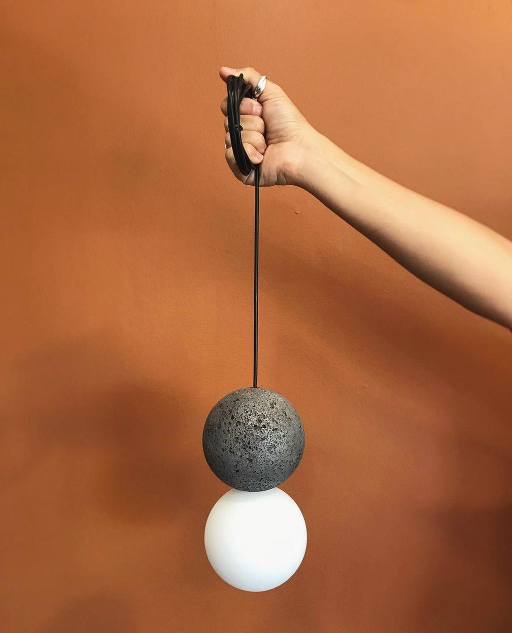 A hand holds this Clérachic pendant by its cord, its dark sphere and opal glass globe set against a rust-orange wall.