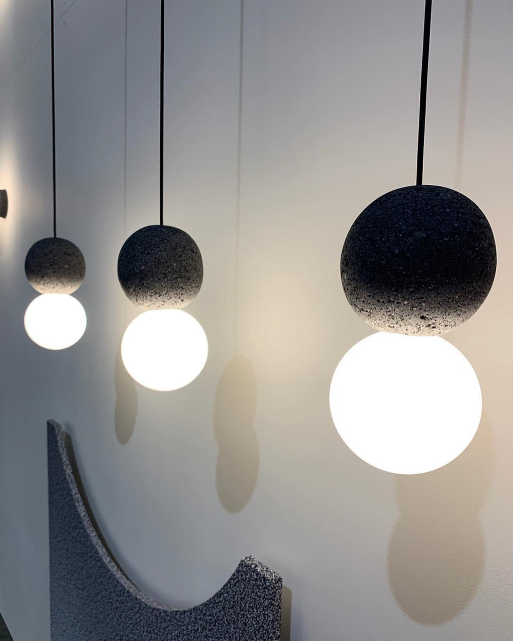 Three Clérachic pendants of different sizes hang together, their dark spheres and opal glass globes lit above a curved grey headboard.