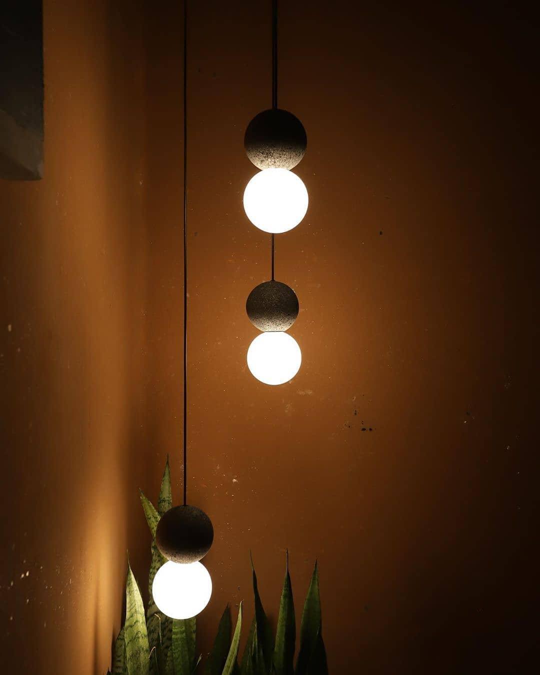 Two Clérachic pendants glow at different heights against a warm, dimly lit amber wall with a plant below.