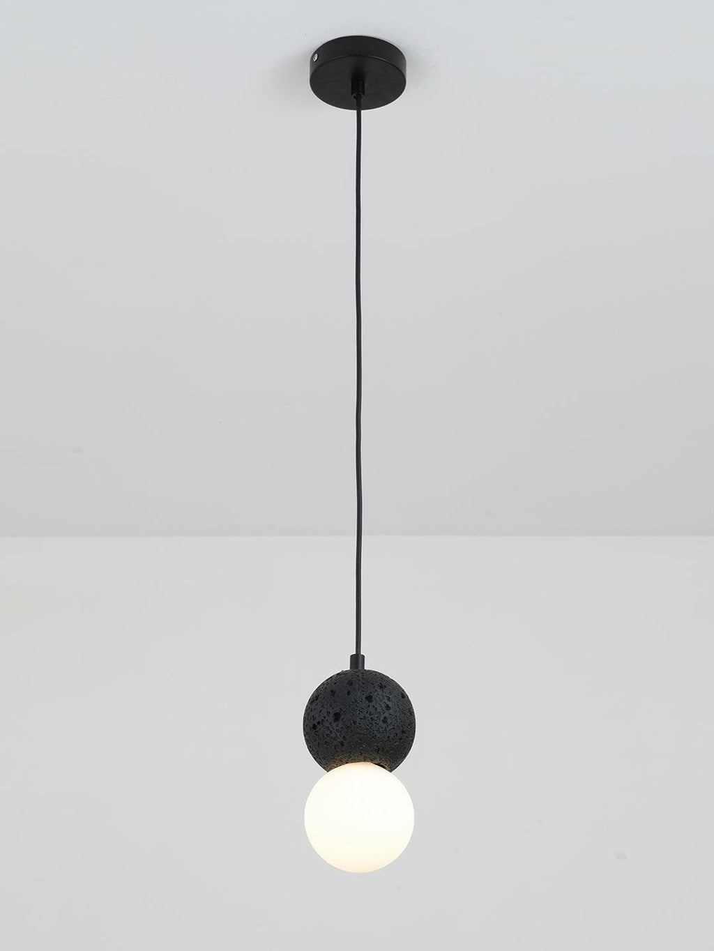This Clérachic pendant, a dark textured sphere above an opal glass globe, drops from a plain white ceiling on a black cord.