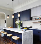 Clérachic pendants line a kitchen island with marble countertop, navy cabinetry and wooden bar stools.