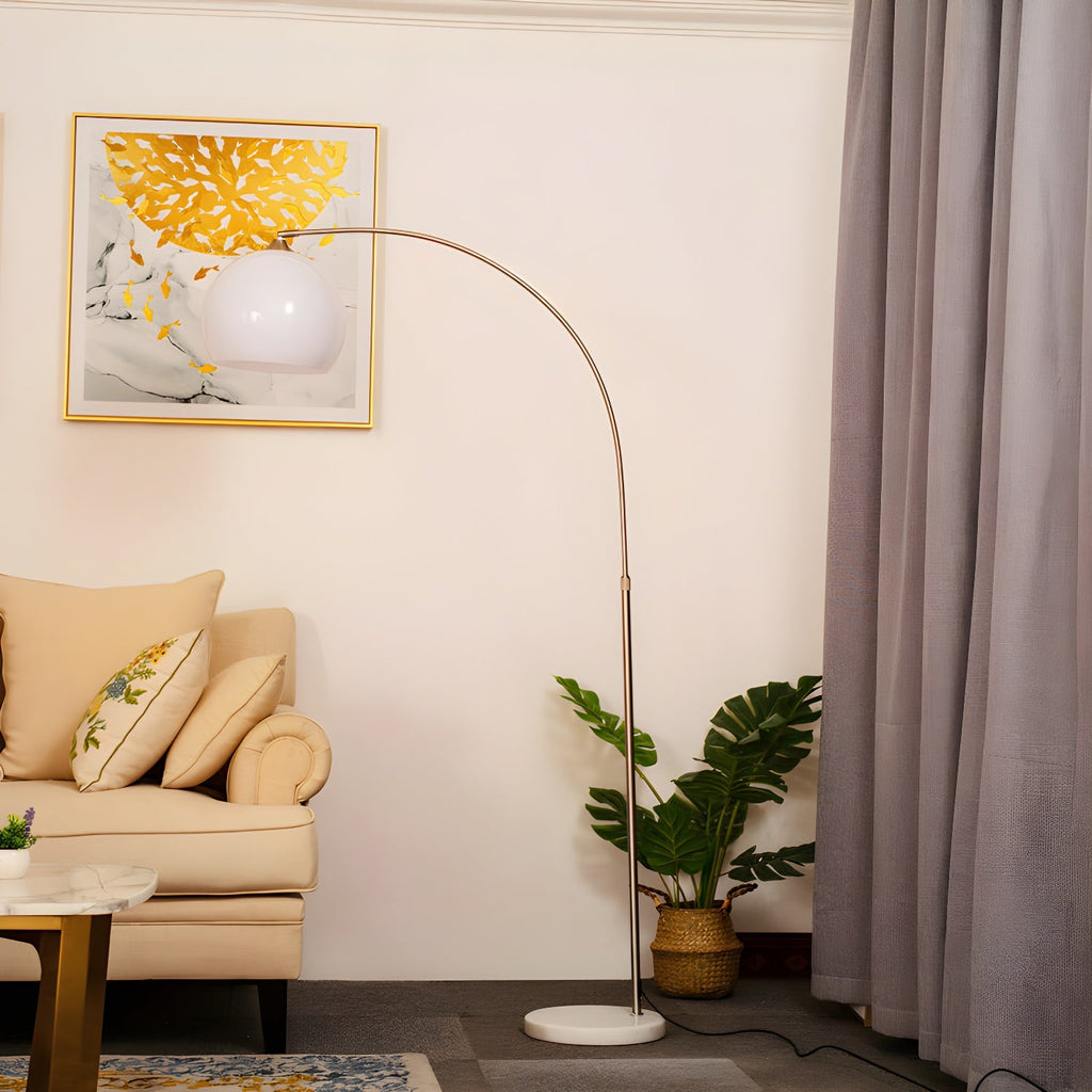 A Clérachic arc floor lamp with a white dome shade curves over a cream armchair near a window with mauve curtains.