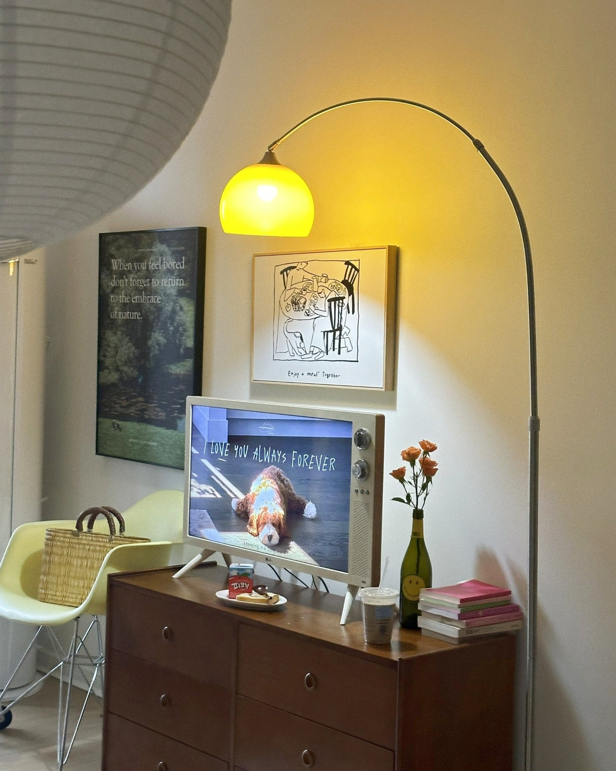 A Clérachic arc floor lamp glows warm yellow above the same dresser and television, a paper lantern visible in the corner.