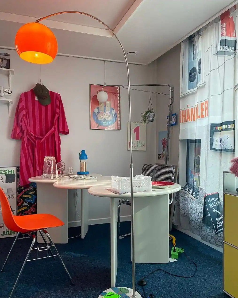 An orange-shaded Clérachic arc floor lamp stands over a white round table in a home office, a red robe hanging on the wall.
