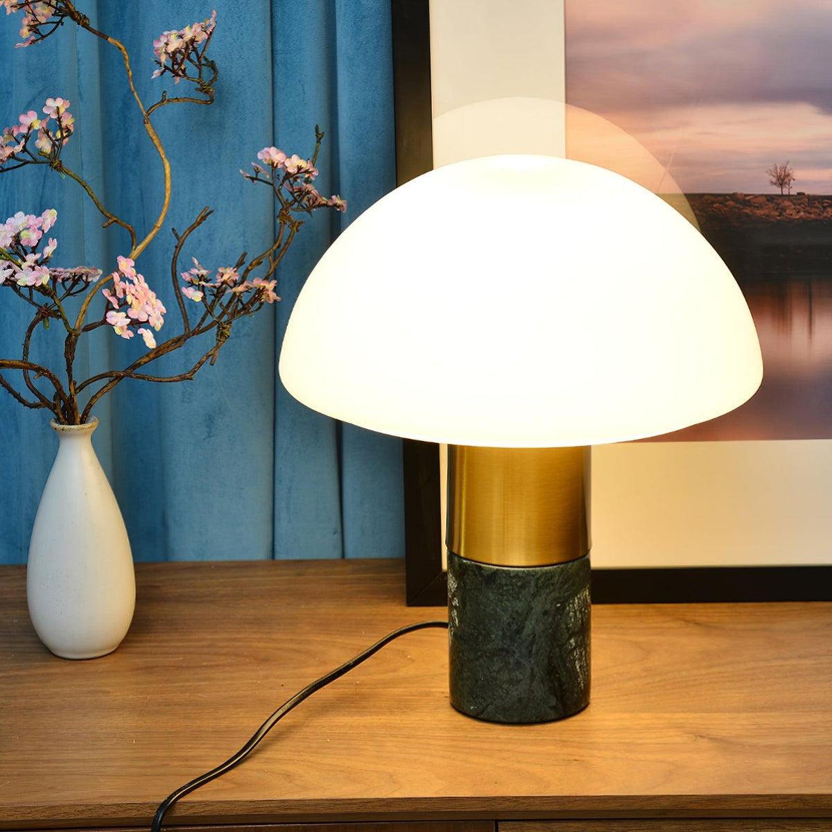 On a wood console styled with pink blossom branches, a Clérachic domed table lamp with a green marble base glows by a teal curtain.