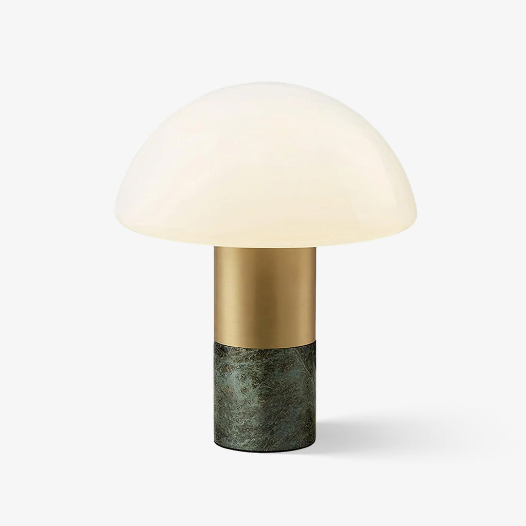 A Clérachic domed glass table lamp with a brushed gold stem and green marble base glows softly on a plain grey backdrop.