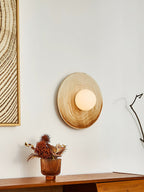 A lit Clérachic ceramic circle sconce seen from across a room, mounted above a walnut sideboard beside framed wall art.