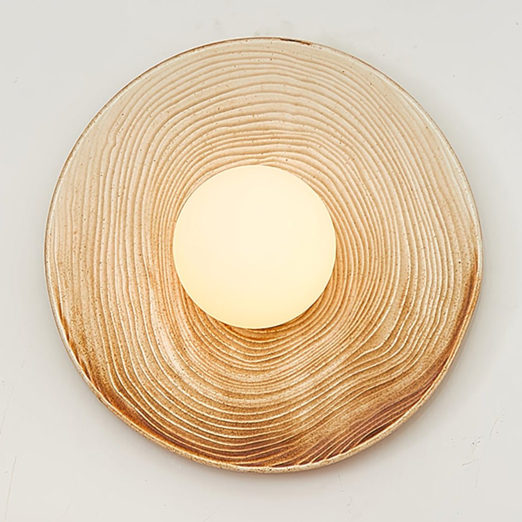 Close-up of a Clérachic ceramic circle sconce, its ridged terra-beige surface radiating outward from the white opal globe.