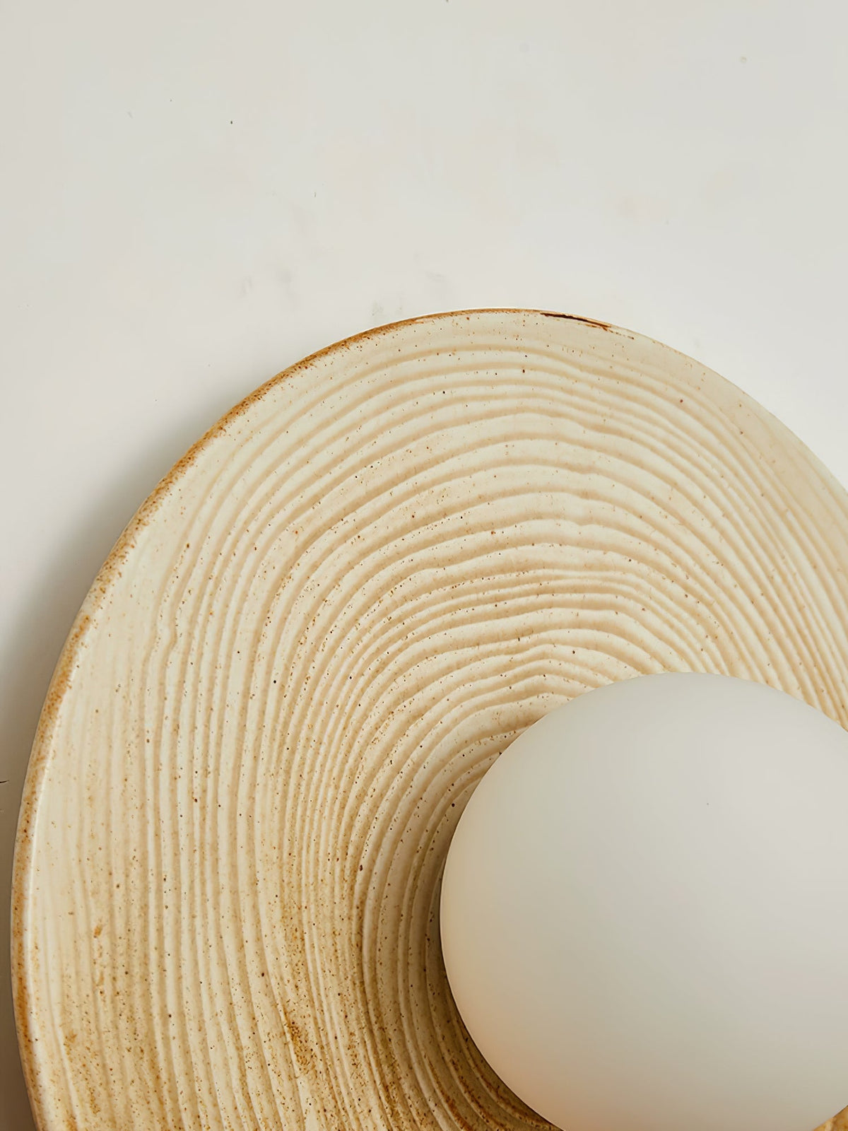 Close-up of the ridged terra-beige ceramic disc on a Clérachic wall sconce, its opal globe set low against the grooved texture.